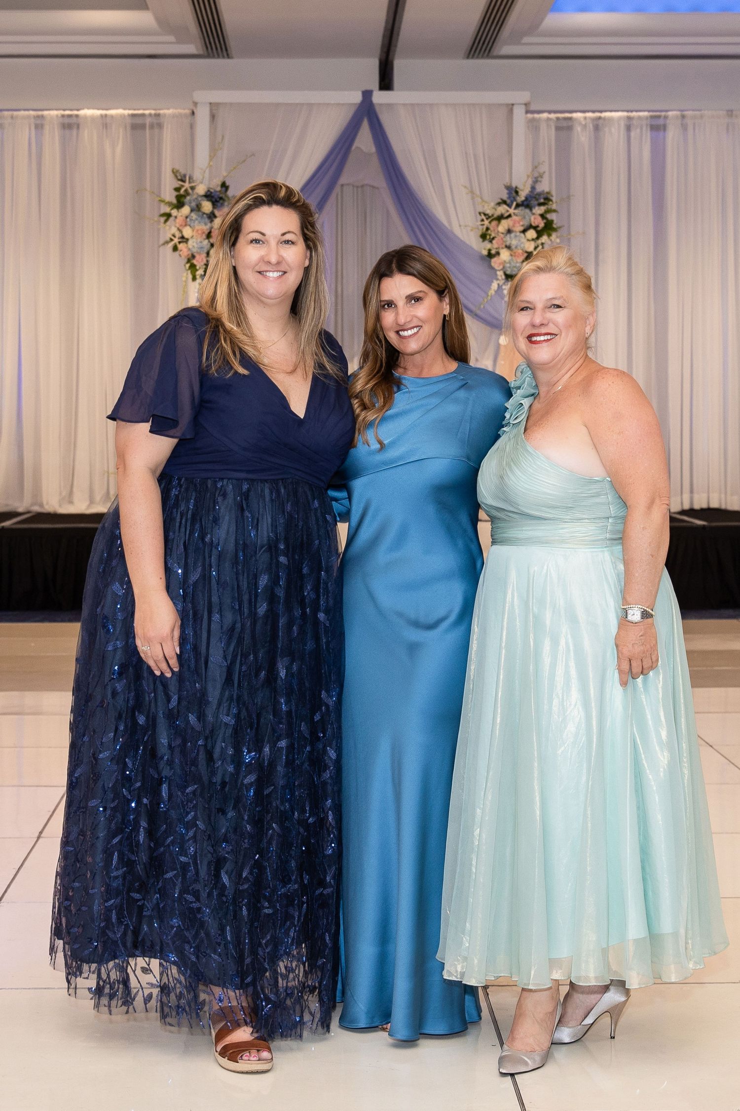Three women in elegant blue gowns pose together at a formal event with floral decorations in the background.
