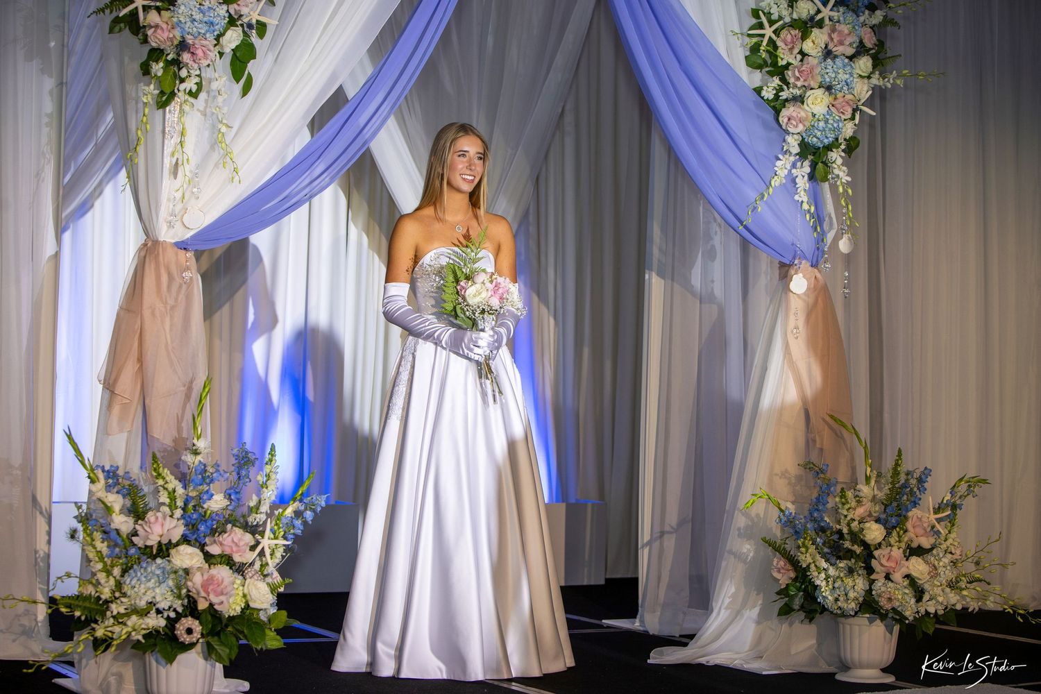 Elegant bride in strapless white gown holding bouquet stands under floral arch with blue draping.