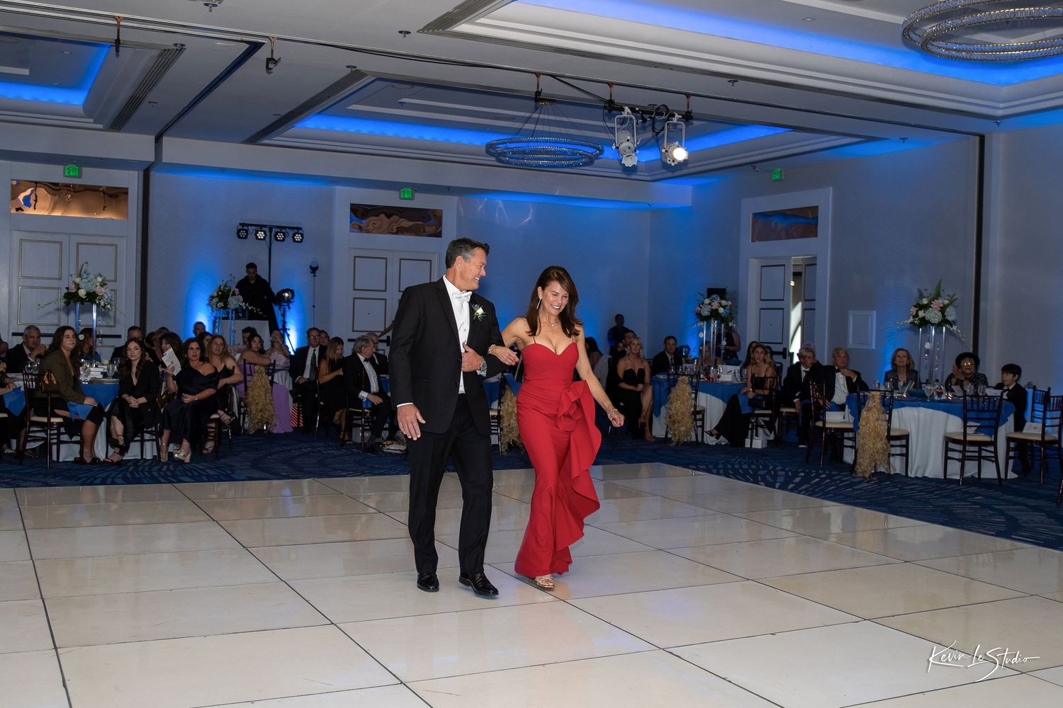 Couple in formal attire dancing on white dance floor at elegant wedding reception with blue lighting.