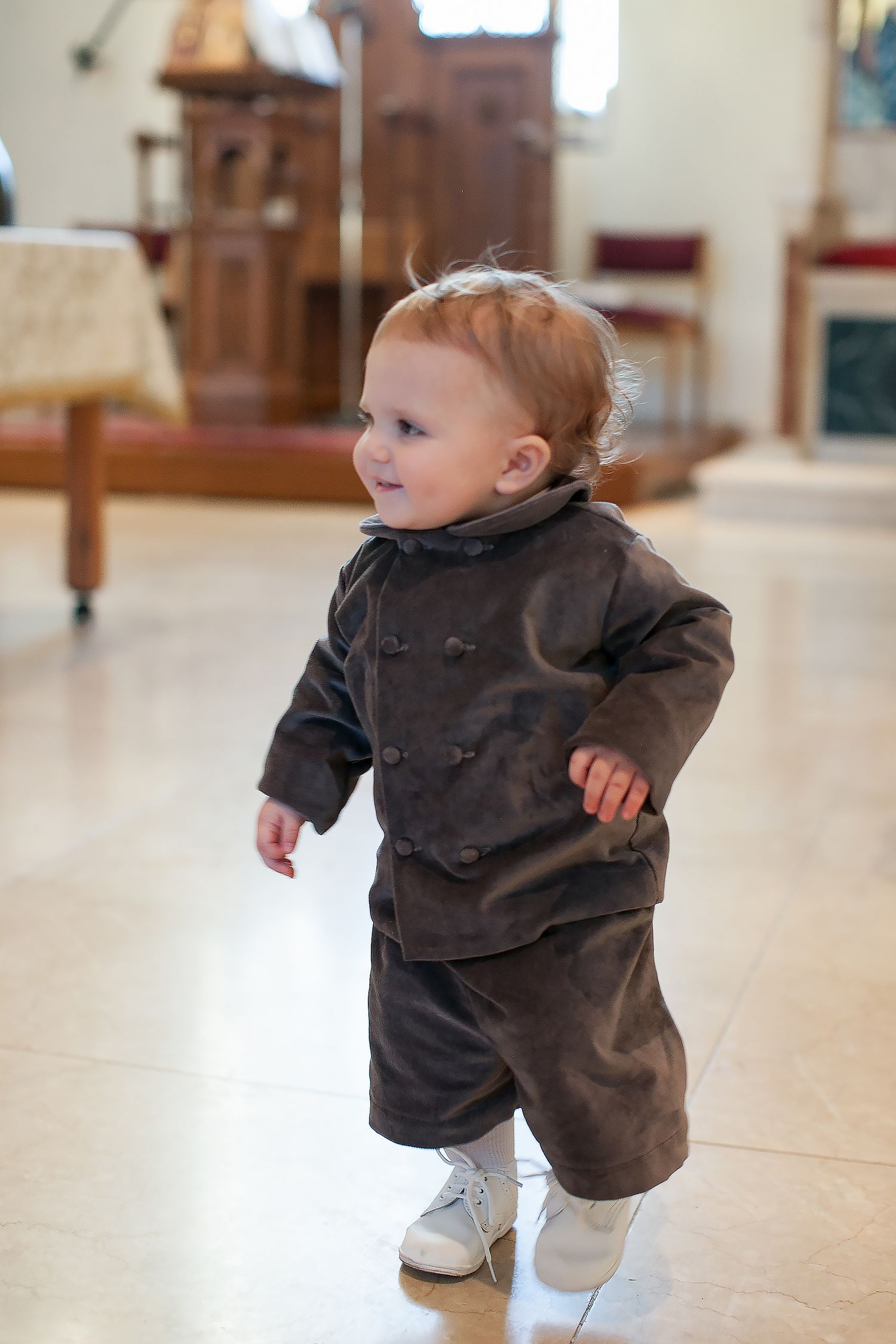 Adorable toddler in brown velvet suit and white shoes standing in a church interior.