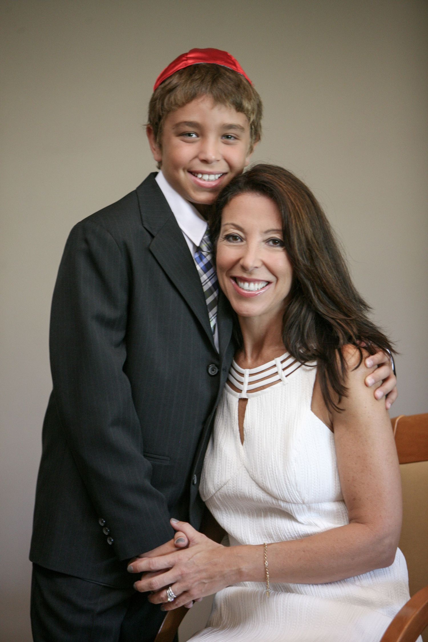 A smiling boy in a suit and red kippah poses with a woman in white dress at what appears to be a Bar Mitzvah.