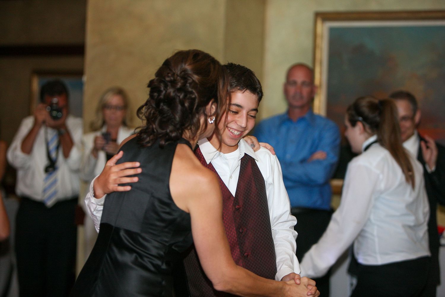 A young boy in a white shirt and vest shares a dance with a woman in black at a formal celebration.