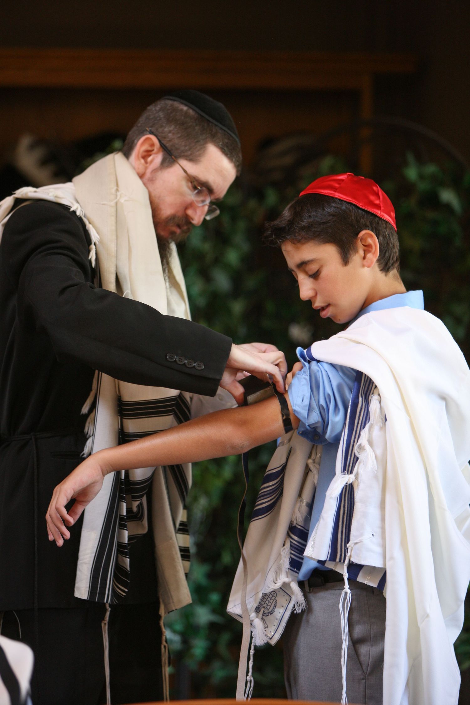 Rabbi helps young boy with tallit during Bar Mitzvah ceremony, both wearing traditional Jewish prayer shawls.