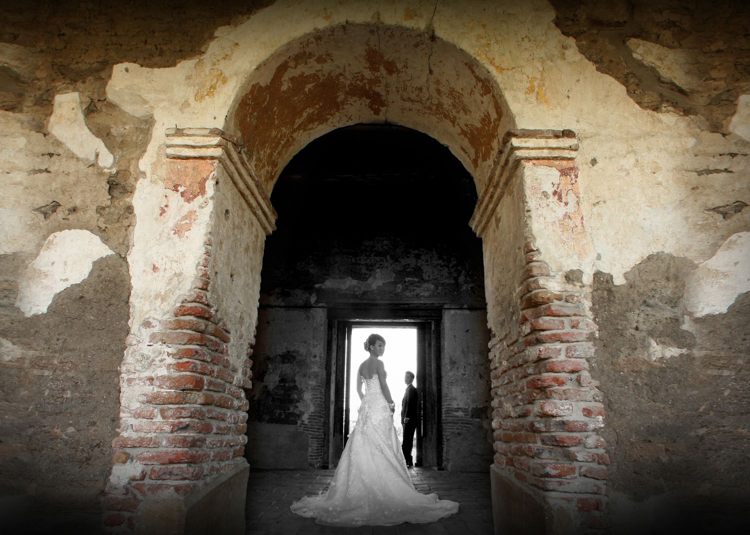 Bride and groom silhouetted in an ornate brick archway of a rustic historic building during a romantic wedding ceremony.