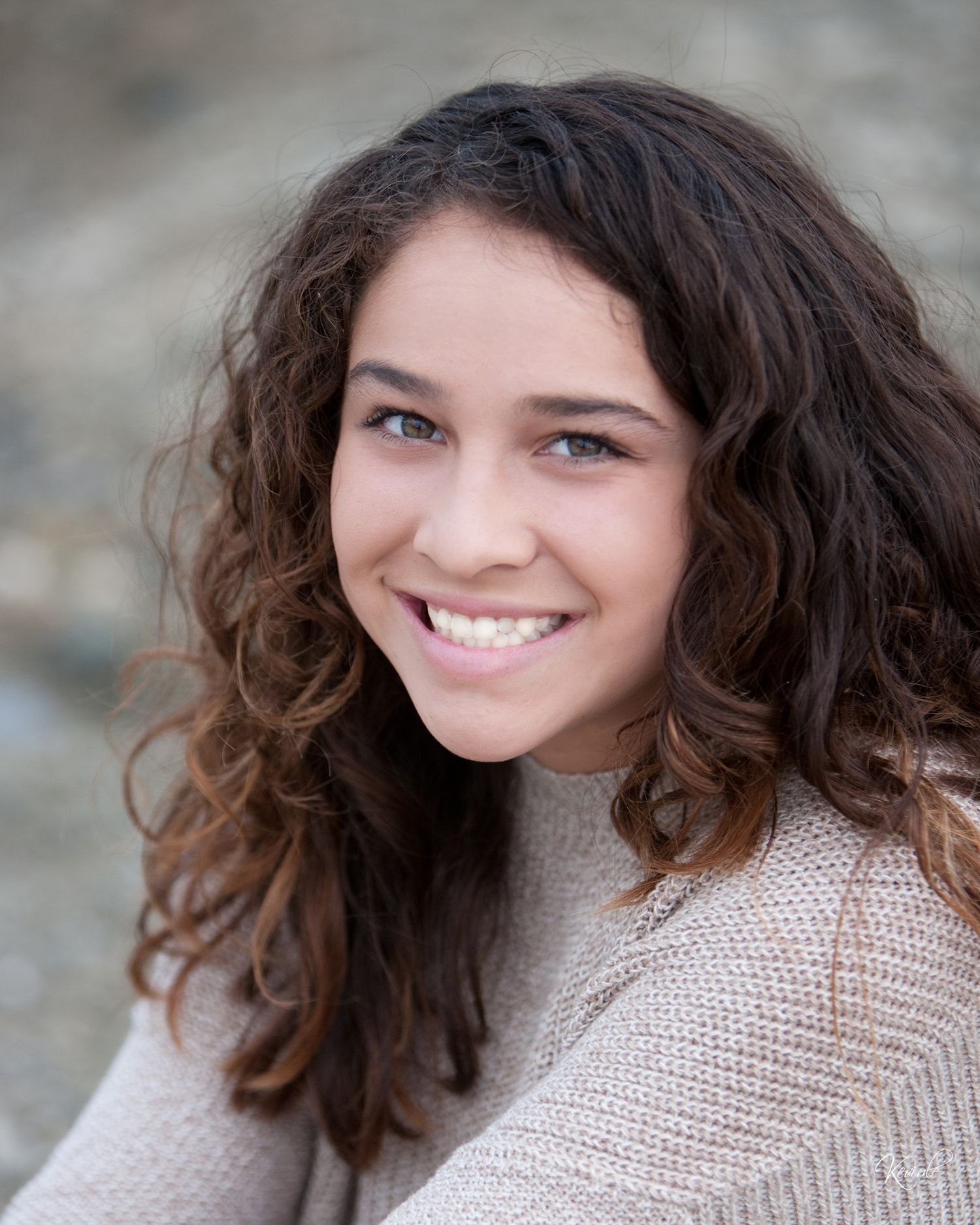 Young woman with curly brown hair and blue eyes smiling broadly in an outdoor professional headshot.