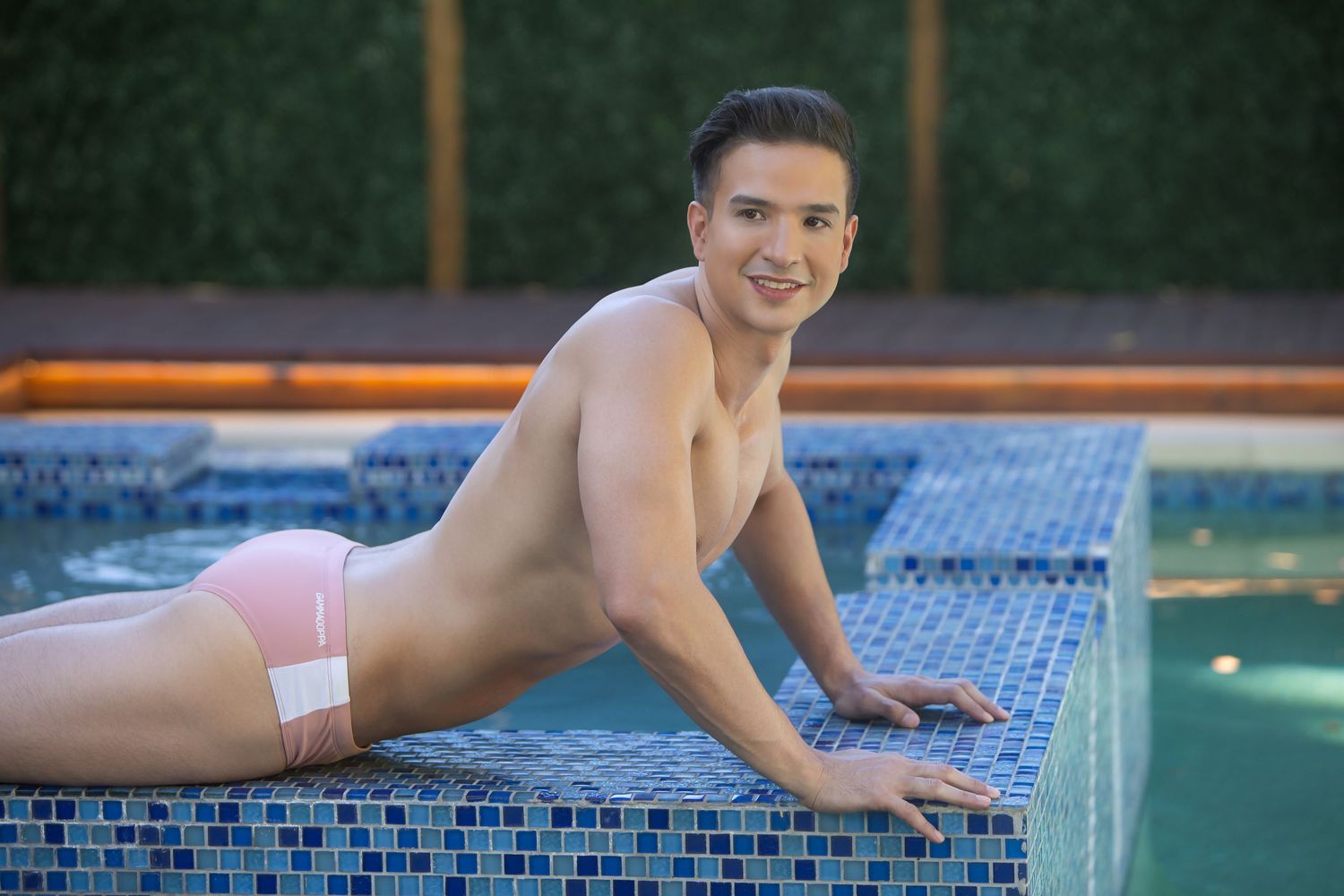 Smiling shirtless young man in pink swim briefs posing at a mosaic tiled pool.
