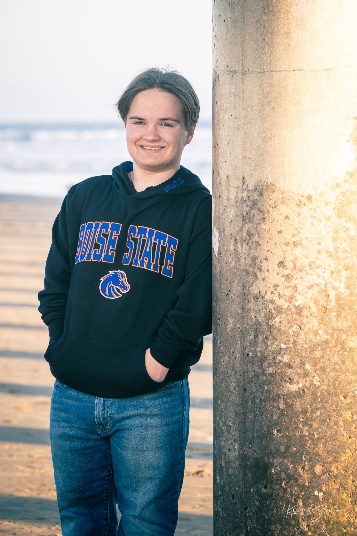 Smiling teen in Boise State hoodie leaning on pier column at beach.