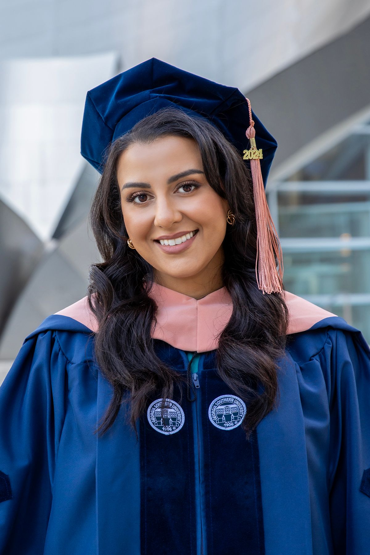 Young woman smiling in navy blue doctoral graduation gown and cap with pink hood and gold tassel.
