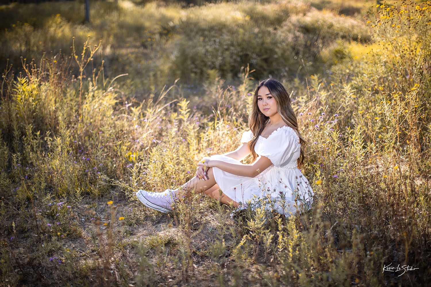 Young brunette woman in white dress sitting relaxed among tall dry grasses in sunny field.