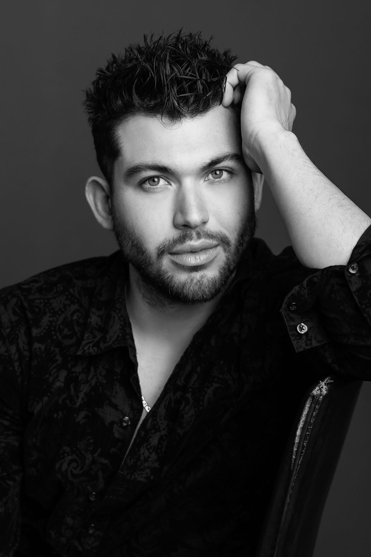 Black and white portrait of a young bearded man in dark patterned shirt with hand in hair, studio shot.