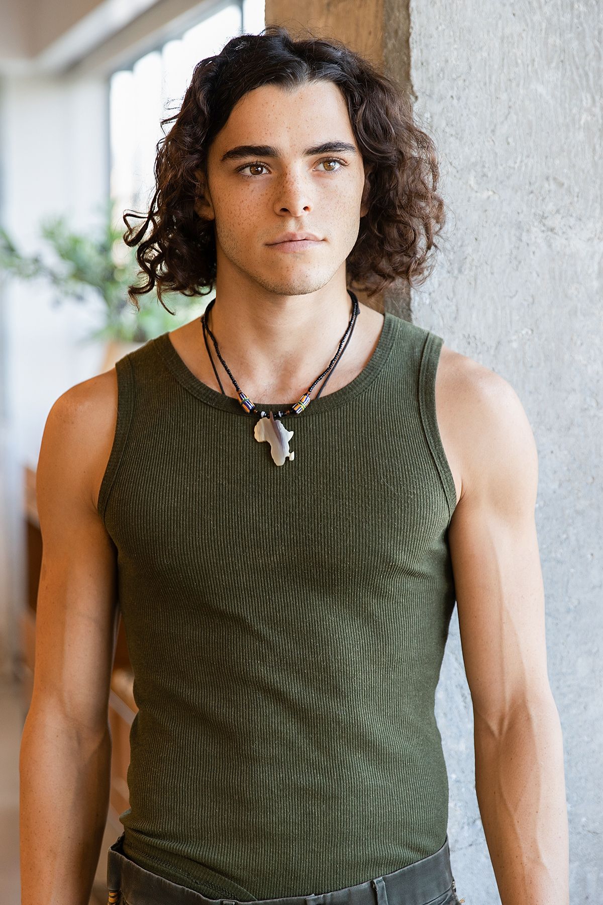Young man with long curly dark hair wearing a green ribbed tank top and layered necklaces, leaning against a wall.