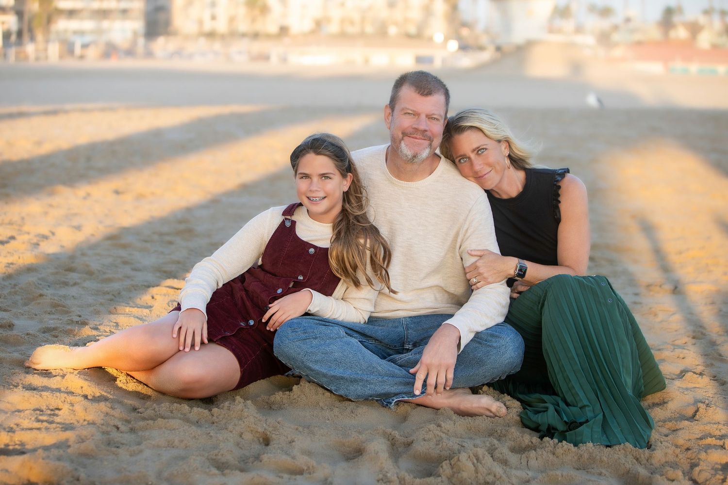 Family of three smiling together on a sandy beach at golden hour, sitting casually in warm sunset light.