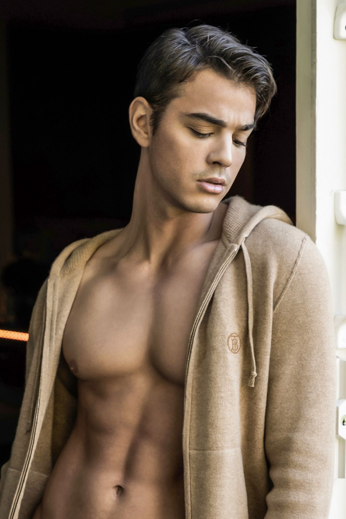 Young athletic man with open beige hoodie showing toned physique, eyes closed, posing in natural light.