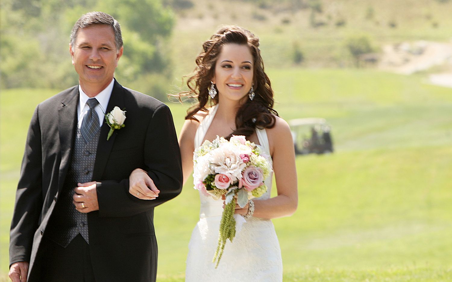Bride in white gown holding pink bouquet walks with suited man on green golf course during outdoor wedding ceremony.