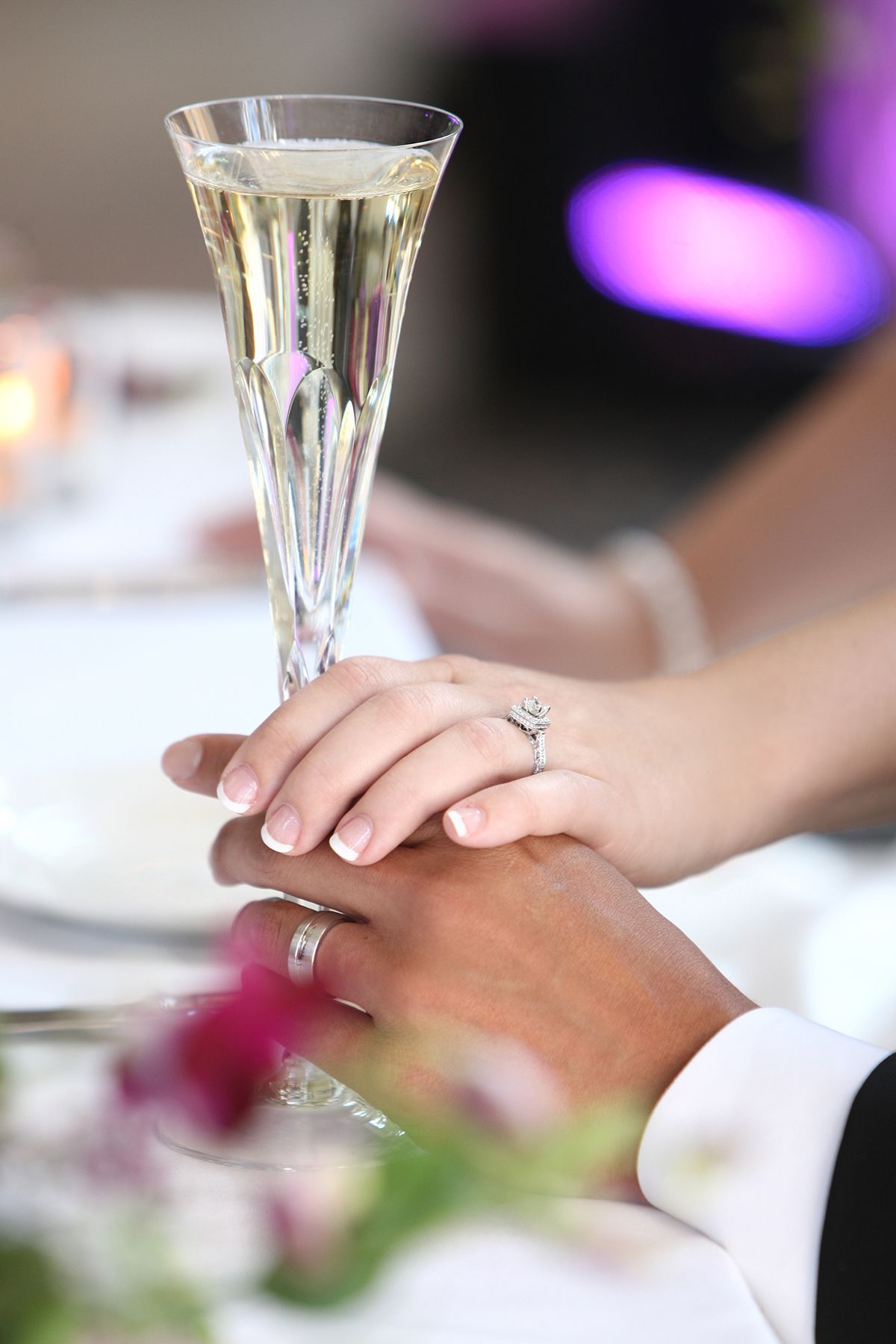 Newlyweds hold hands over champagne flute showing diamond engagement and wedding rings.