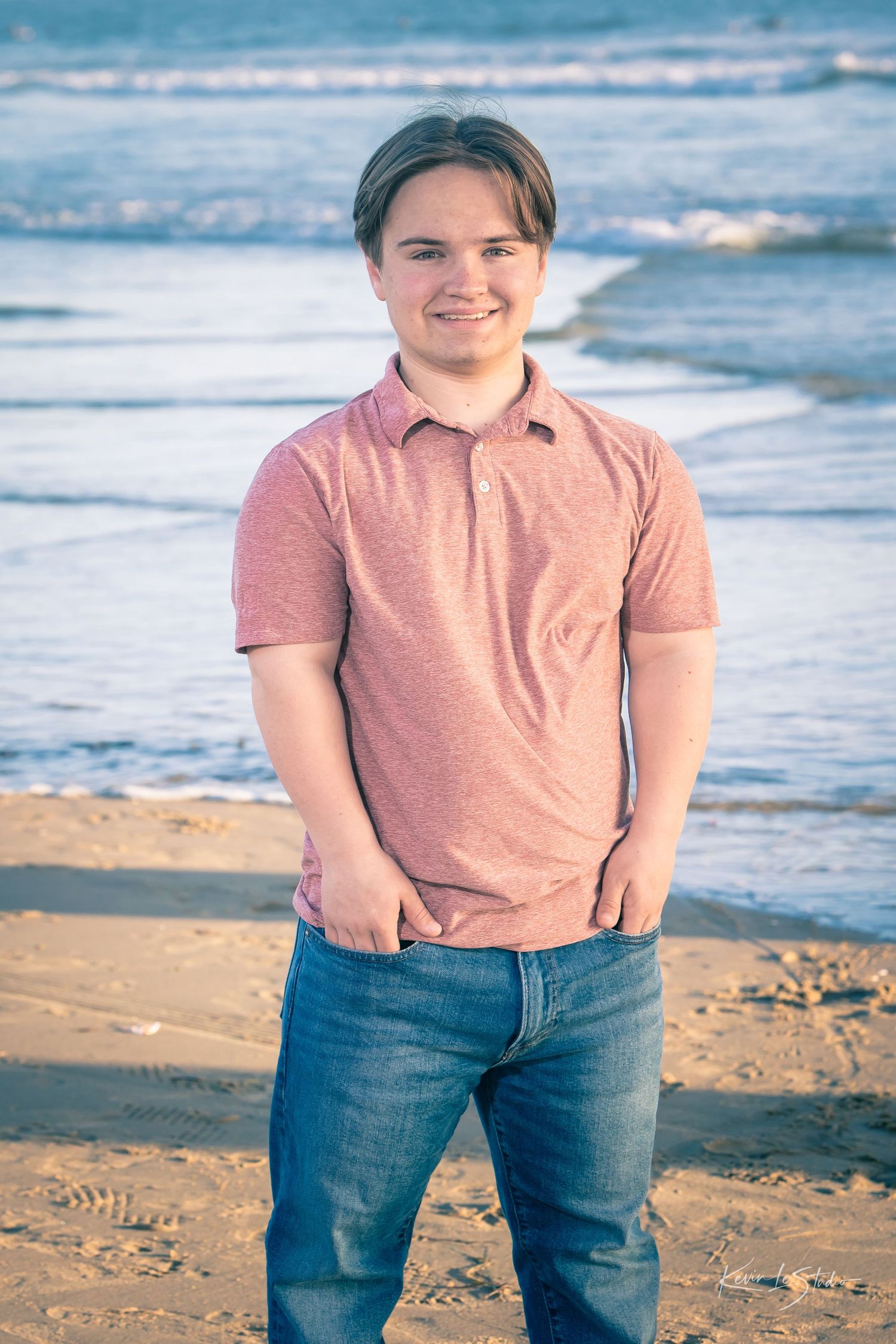 Smiling young man in pink polo and jeans posing on sandy beach.