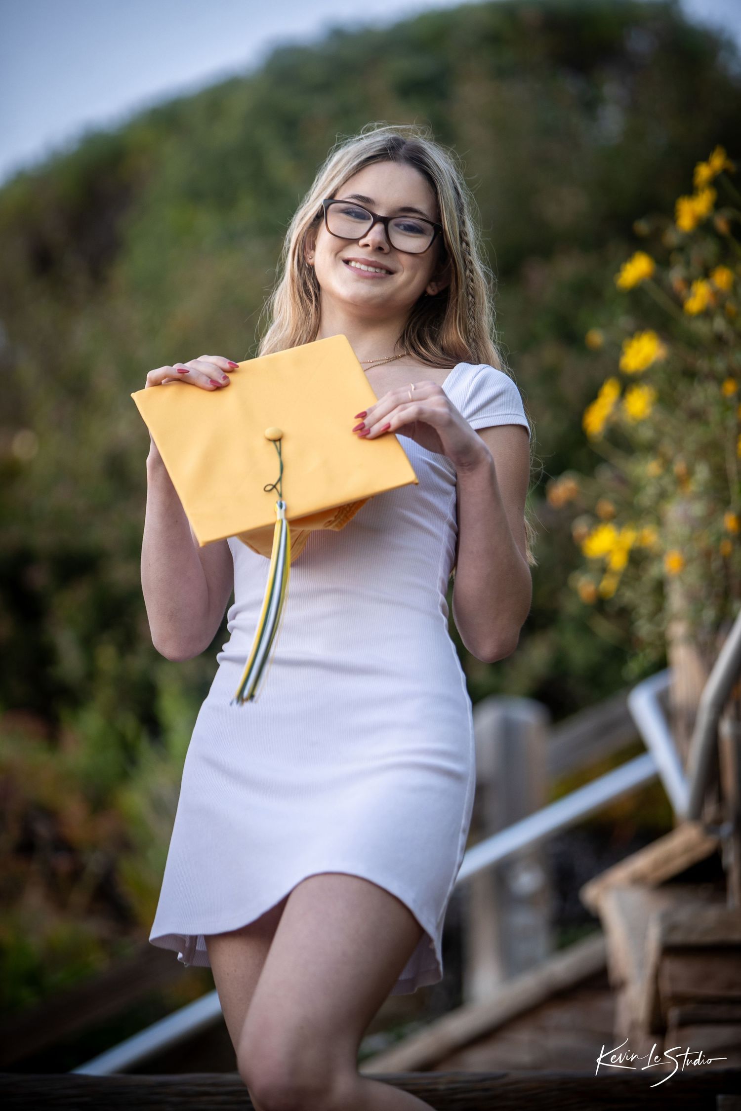 Smiling blonde girl in white dress holding gold graduation cap outdoors with yellow flowers.