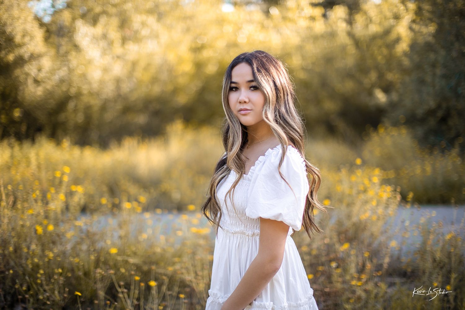 Young woman in white dress poses gracefully in golden field with wildflowers during outdoor senior portrait session.