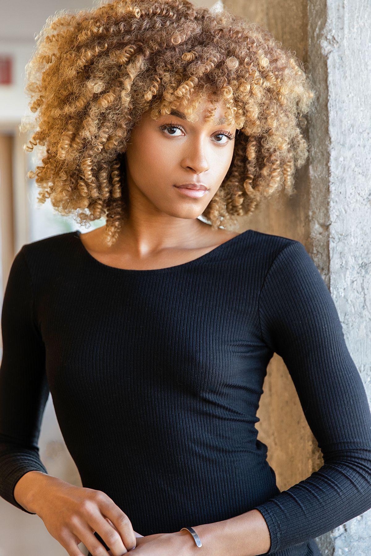 Young Black woman with voluminous blonde curly afro hair wearing a black long-sleeve ribbed top, posing indoors.