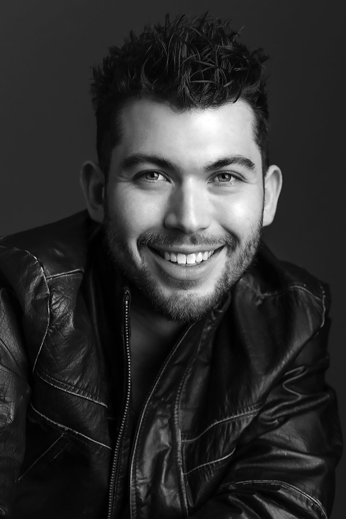 Smiling young man with dark curly hair wearing a leather jacket in a black and white portrait photo.