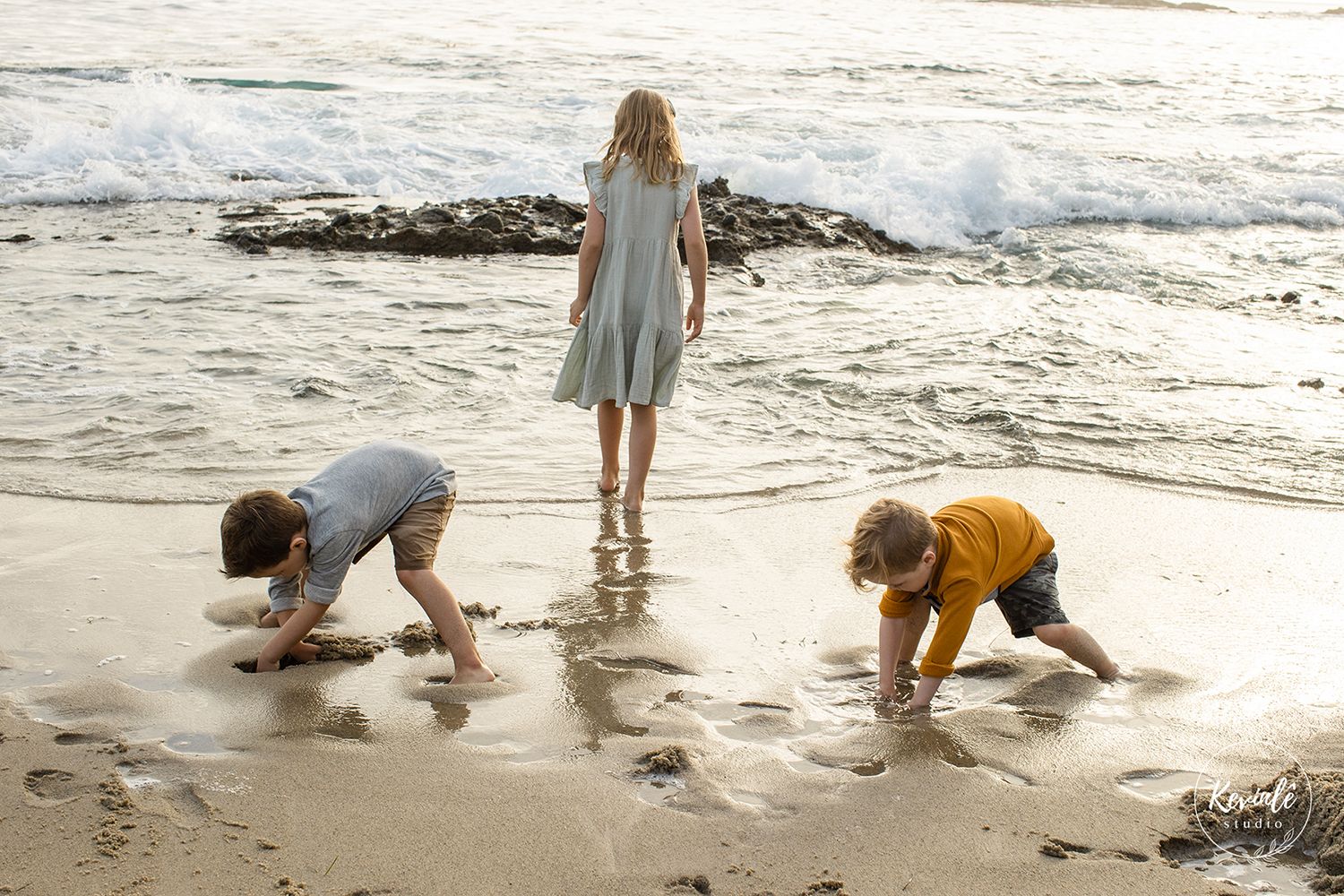 Three children playing on a sandy beach shore, two crouching in wet sand while one girl stands watching ocean waves.