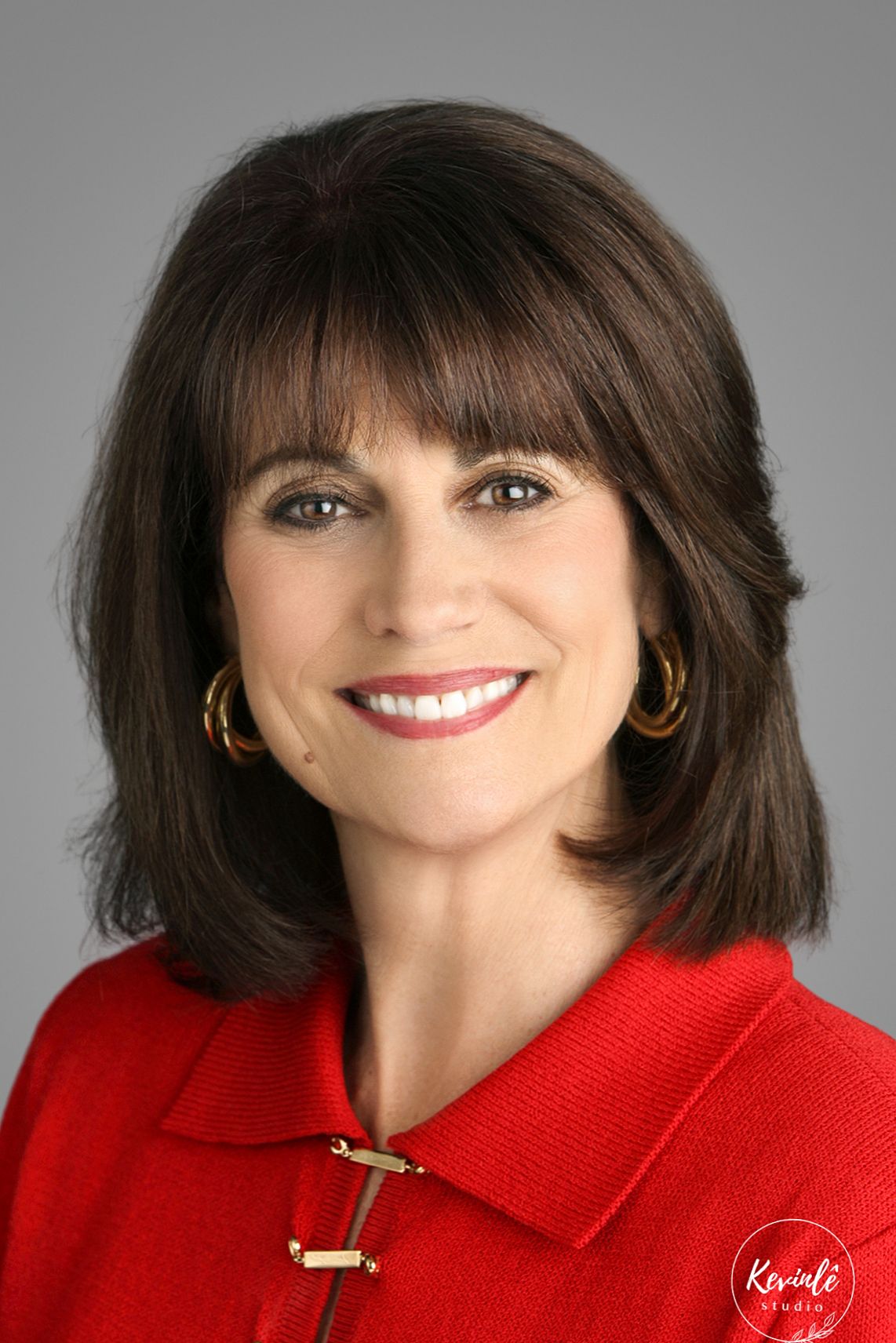 Professional headshot of a smiling woman with dark hair, wearing a red jacket and gold hoop earrings against a gray background.