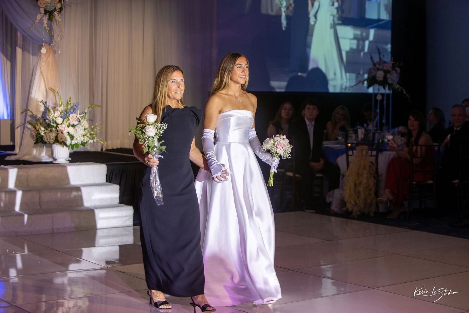 Two elegant women in formal gowns holding bouquets walk a runway at a wedding fashion show.