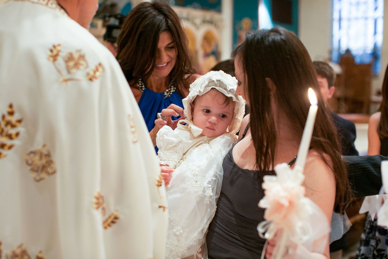 Baby in white christening gown and bonnet held during baptism ceremony with priest and candle.