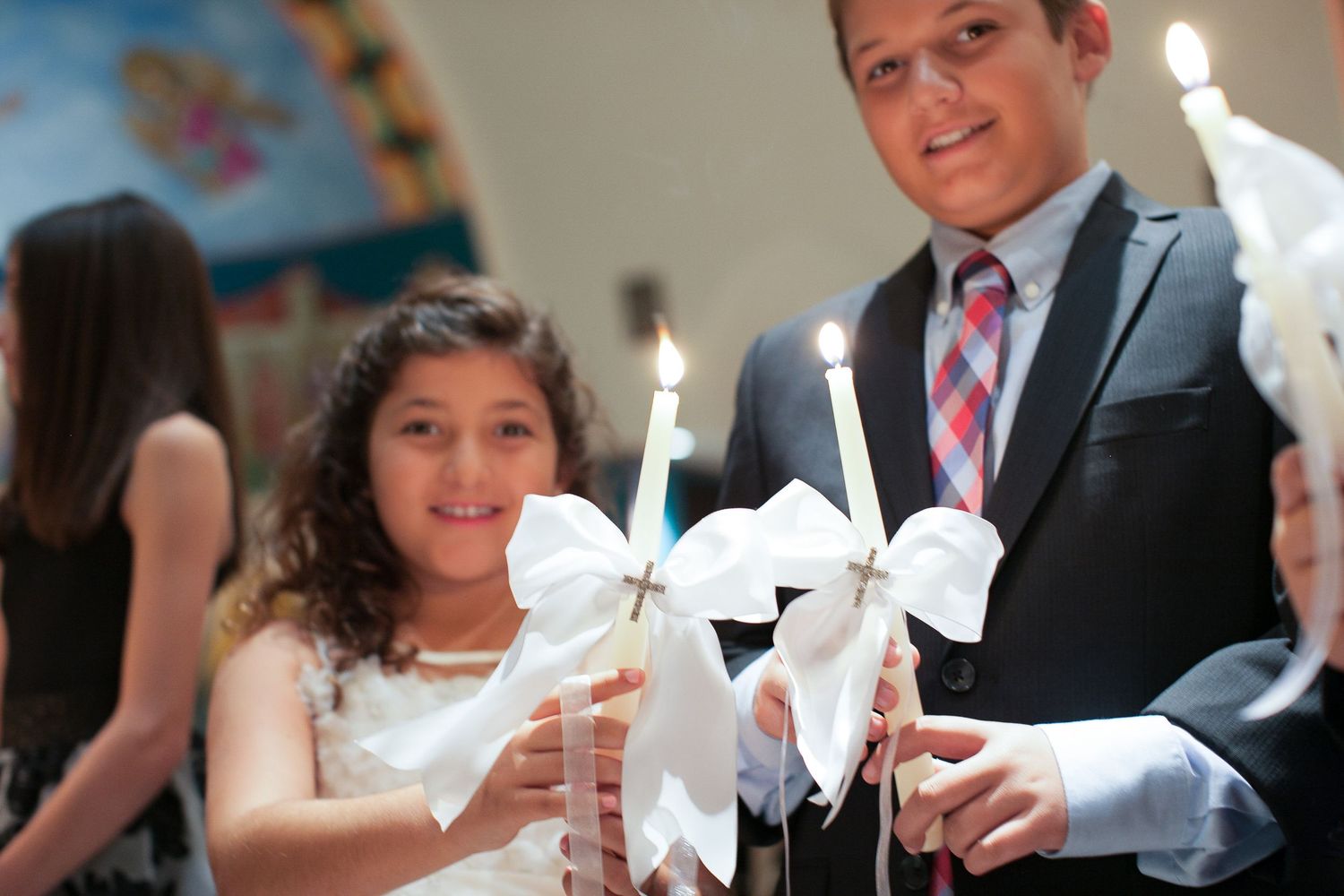 A young girl and teenage boy holding decorated candles with white bows during a religious ceremony.