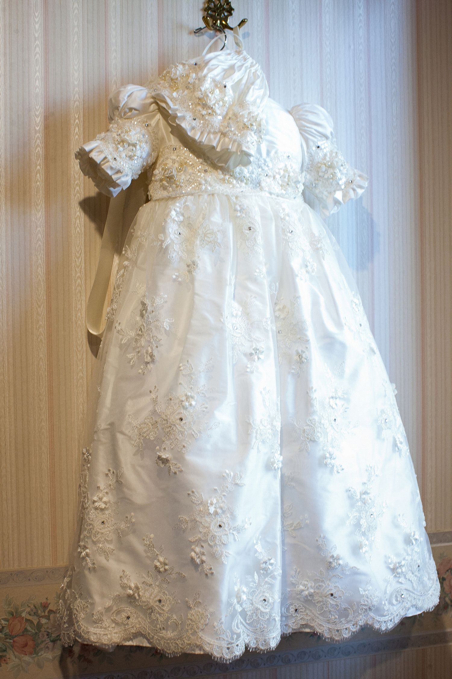 Elegant white baby christening gown with lace embroidery, floral appliques, puff sleeves, and matching bonnet on display.