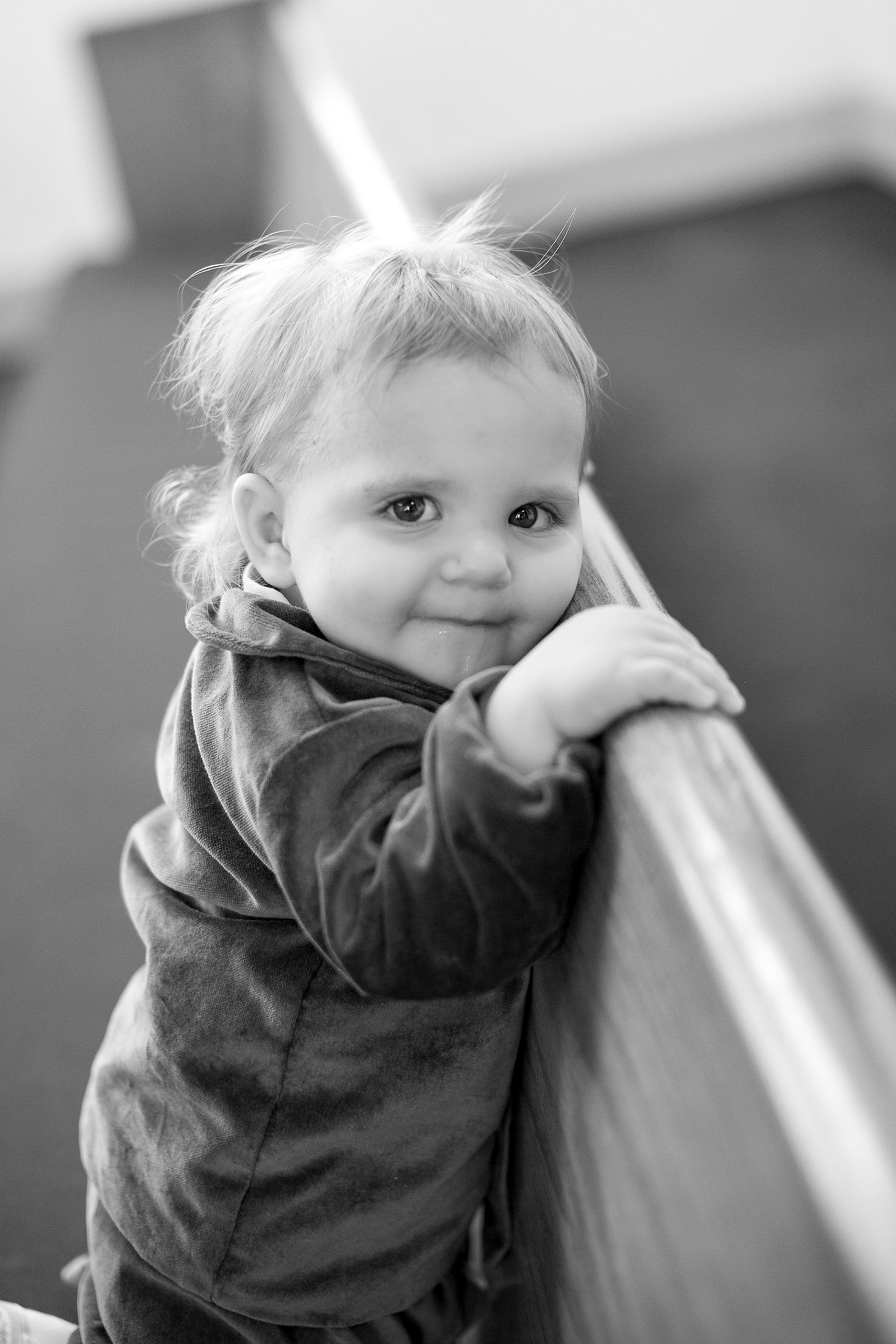 Black and white portrait of a cute toddler leaning on a railing, smiling softly at the camera.
