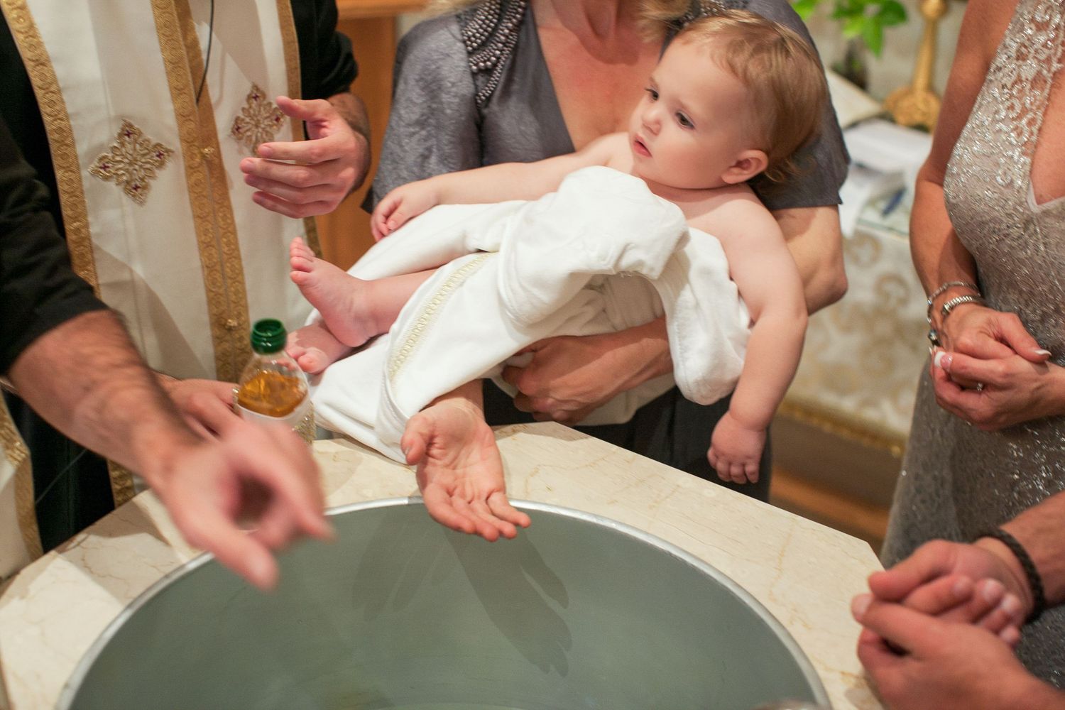Baby in white gown held over baptismal font during Christian christening ceremony with priest and family.