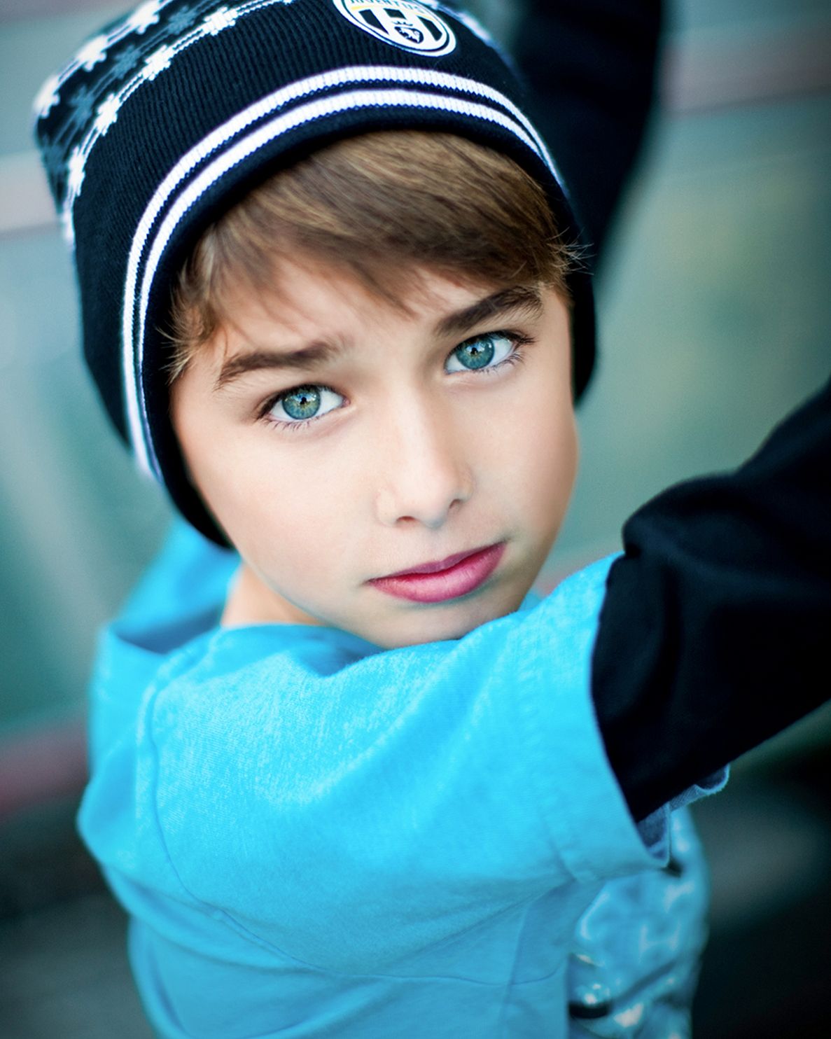 Young boy with striking blue-green eyes wearing a black beanie hat and turquoise jacket in a portrait photo.