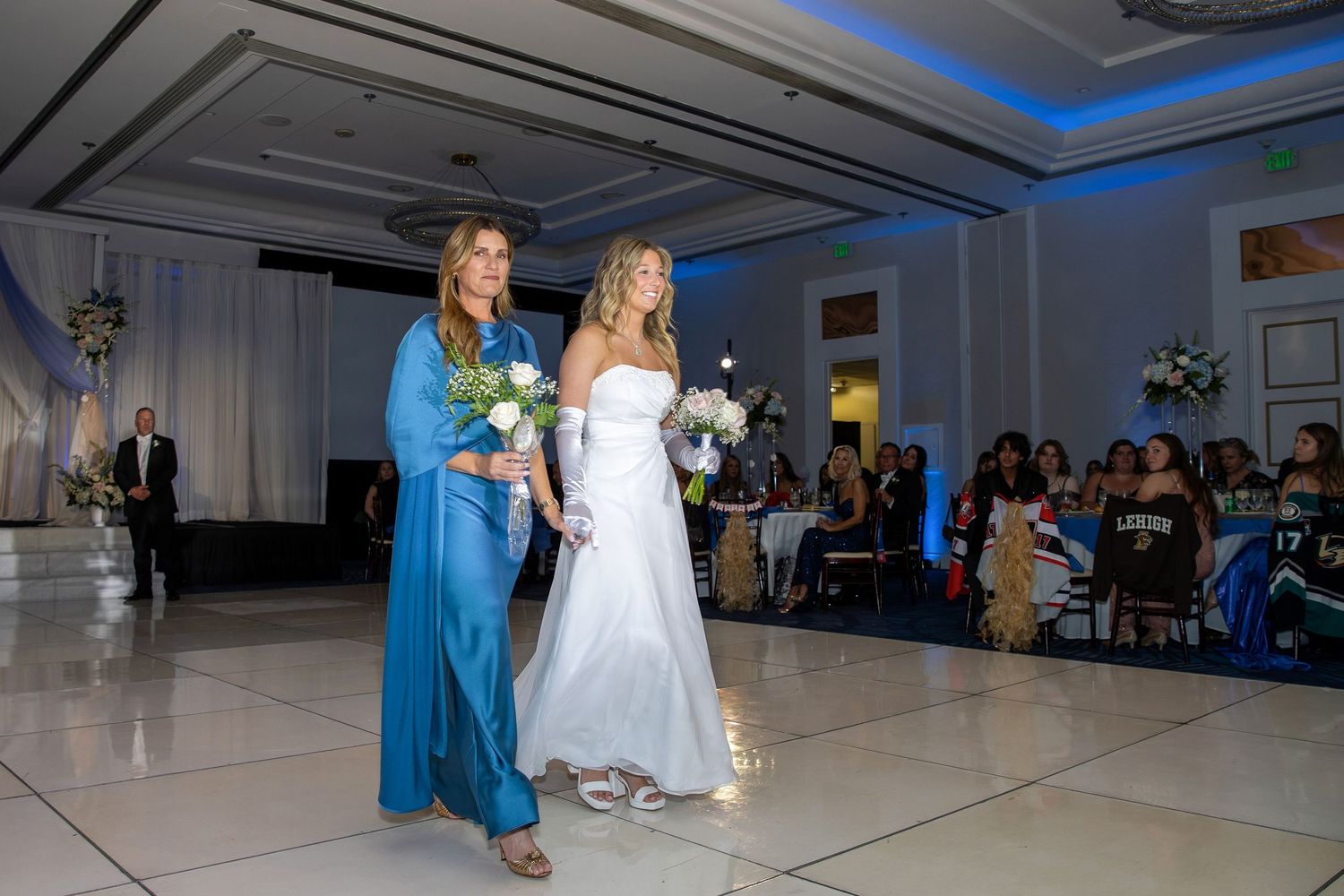 Bride in white gown walks with woman in blue dress holding bouquets at elegant wedding reception venue.