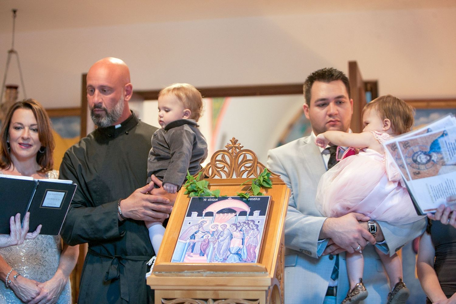 Adults holding babies gather around a framed religious icon during a church baptism ceremony indoors.