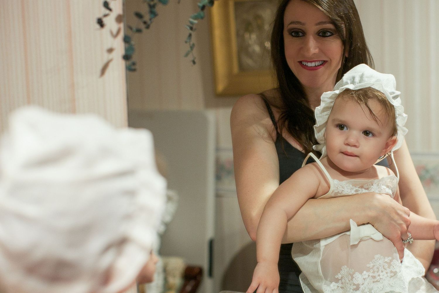 Woman in black dress holding a baby girl wearing a white lace bonnet and dress at a christening event.