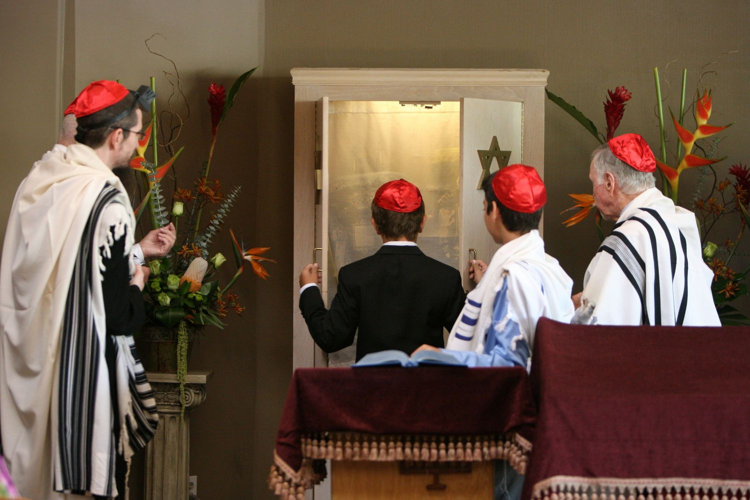 Jewish men in kippot and tallitot pray before an open Torah ark during a synagogue service.