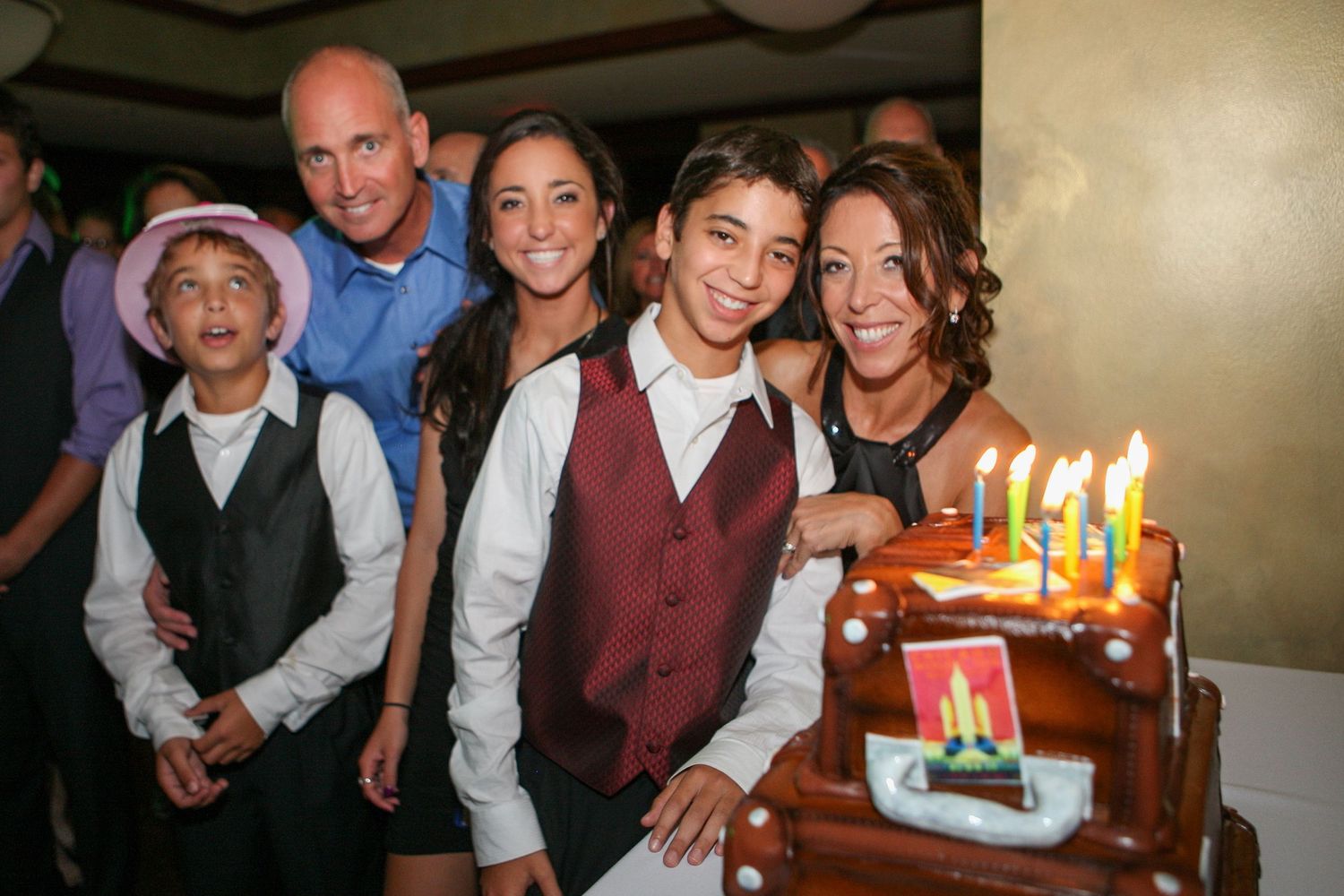 Family posing together smiling behind a multi-tiered birthday cake with lit candles at a celebration.