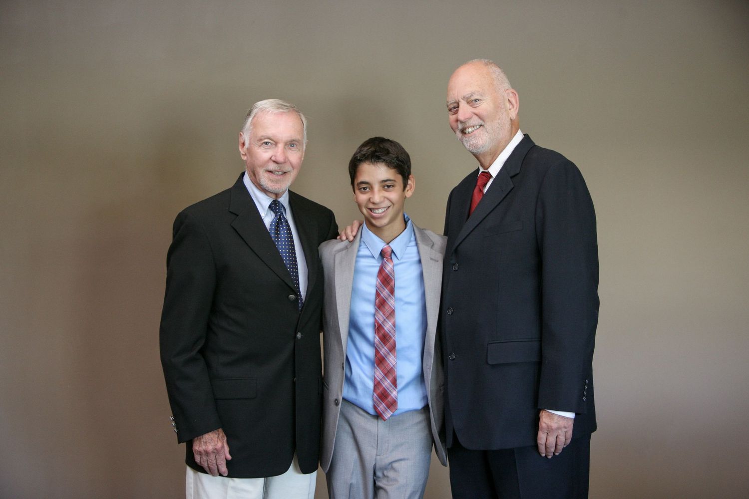 Three men in formal suits smiling together for a portrait photo, with a young teen in the center wearing a blue suit and red tie.