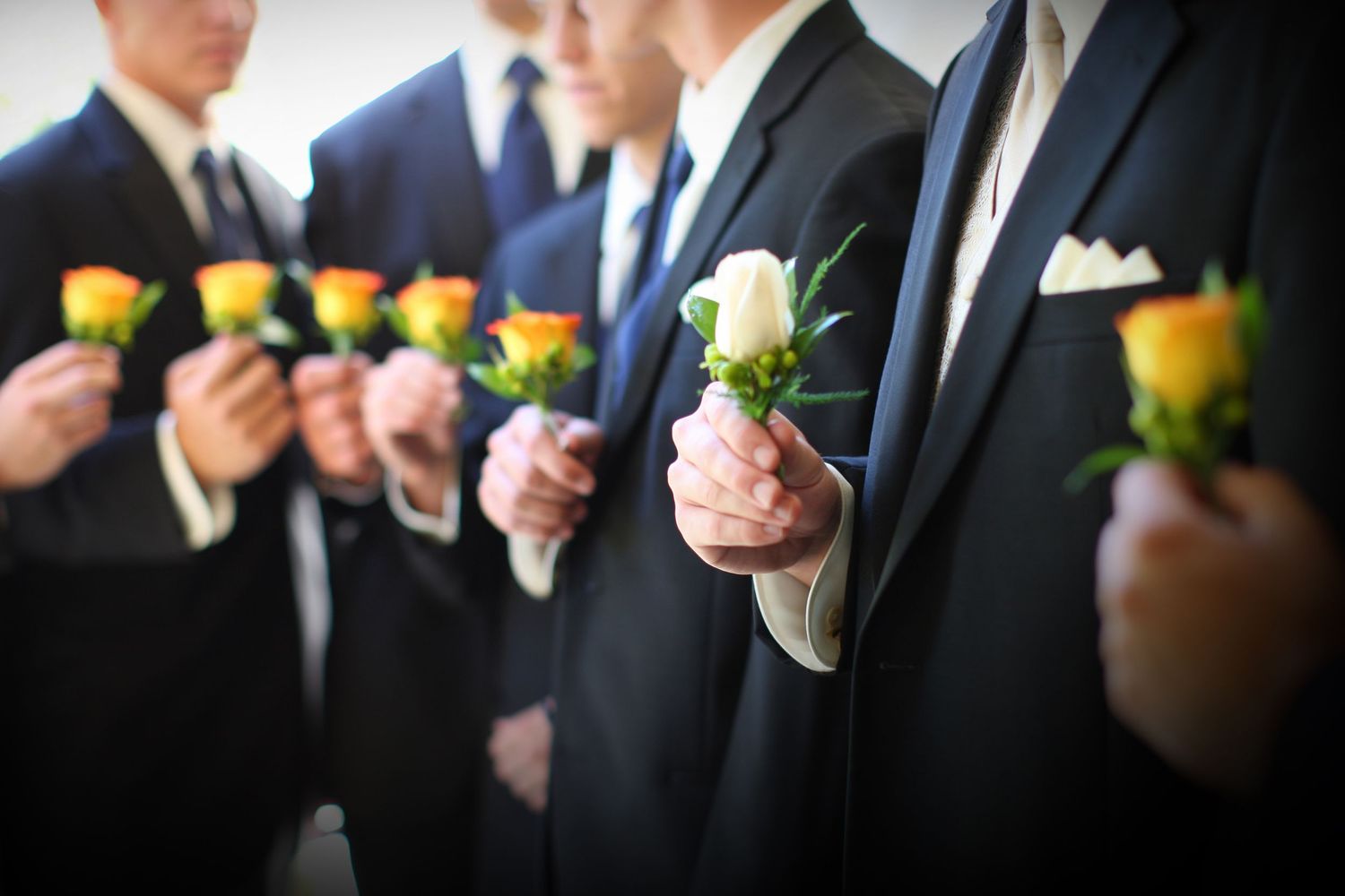 Groomsmen in black suits holding yellow and white rose boutonnieres at a wedding ceremony.