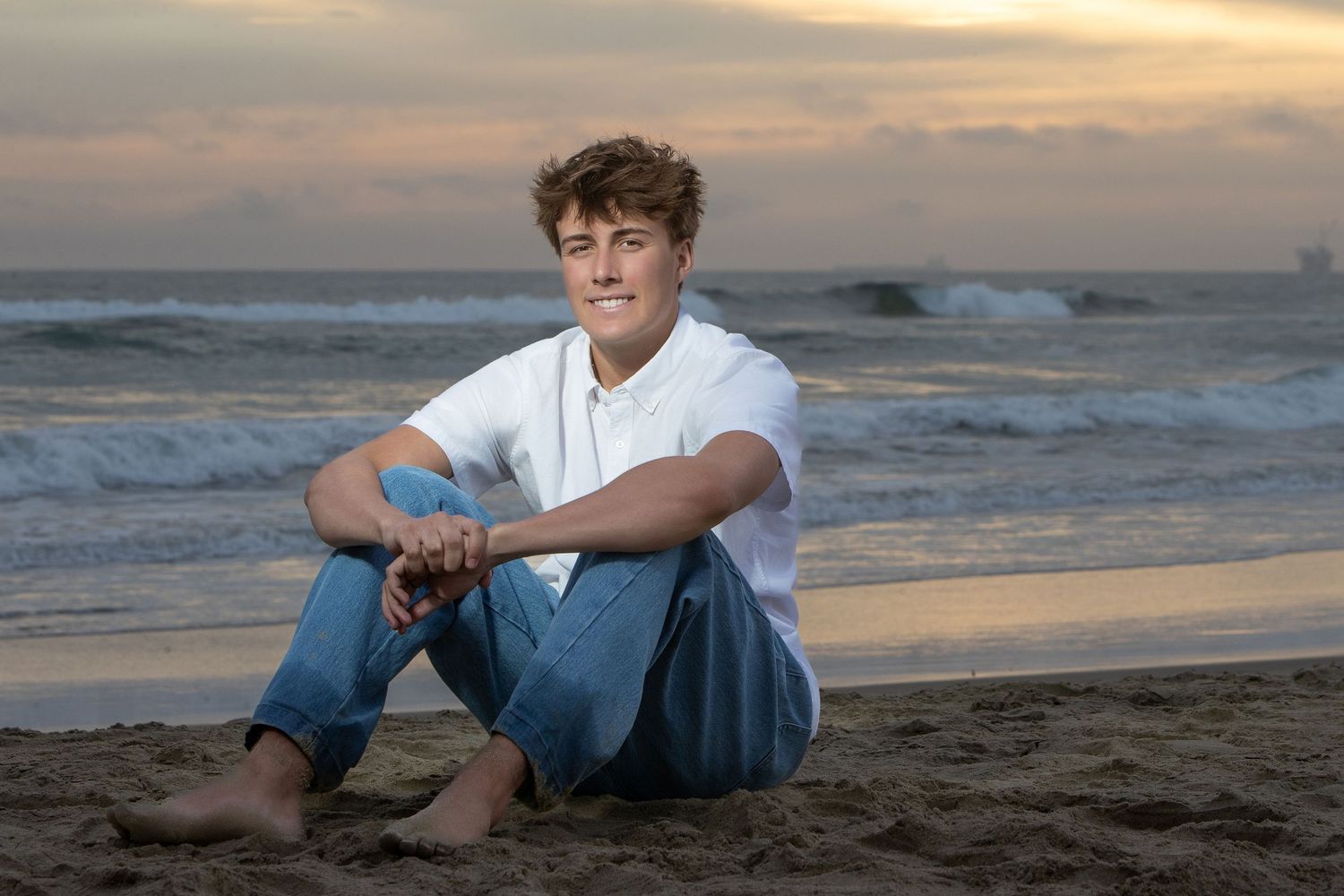 A young man sits barefoot on a sandy beach at sunset, wearing a white shirt and jeans, smiling at the camera.