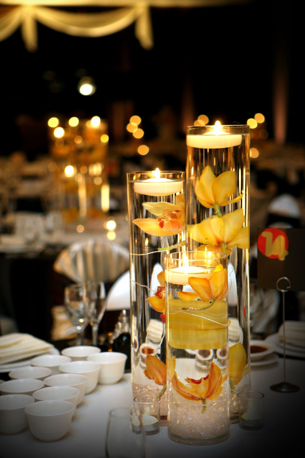 Elegant wedding centerpiece with floating candles and yellow orchids in tall glass cylinders on a reception table.