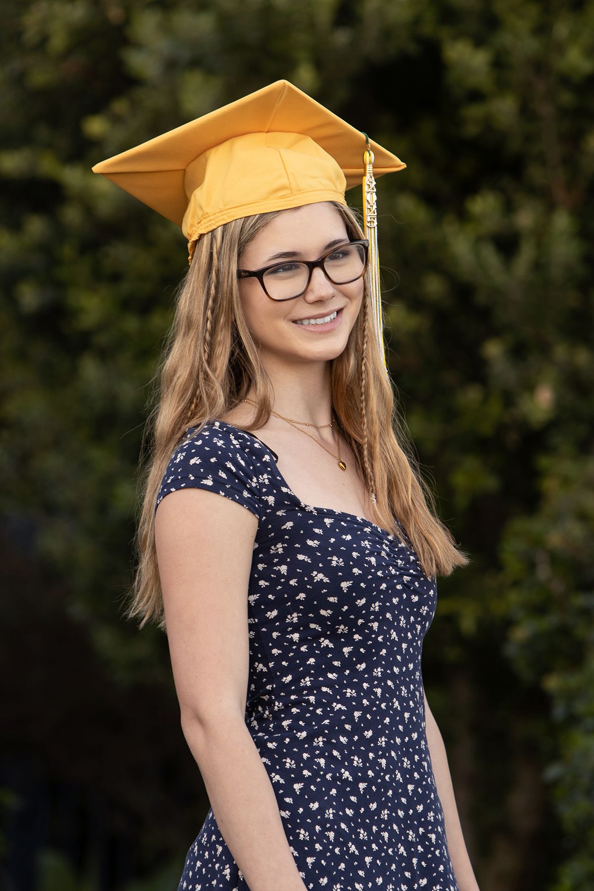 Smiling young woman in gold graduation cap and navy floral dress posing outdoors with green trees behind her.