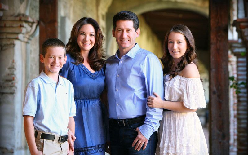 Family of four smiling together in a bright outdoor archway setting for a portrait.