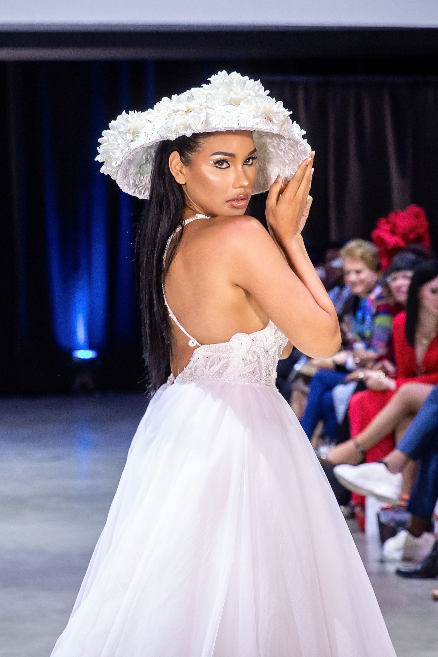 Model in a white floral wide-brim hat and flowing white tulle gown poses on a fashion runway looking over shoulder.