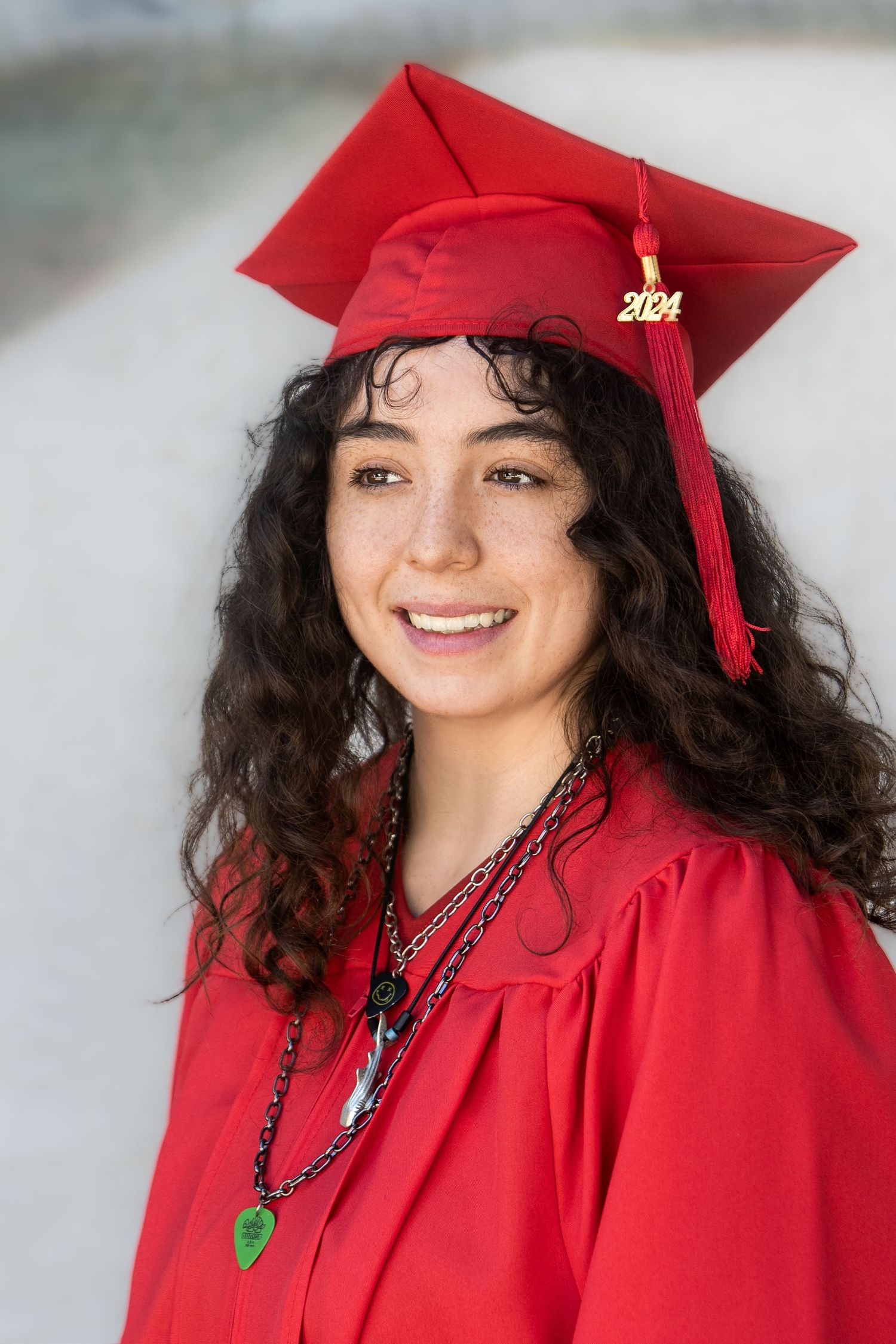 Smiling graduate in red cap and gown with 2024 tassel poses for senior portrait outdoors.