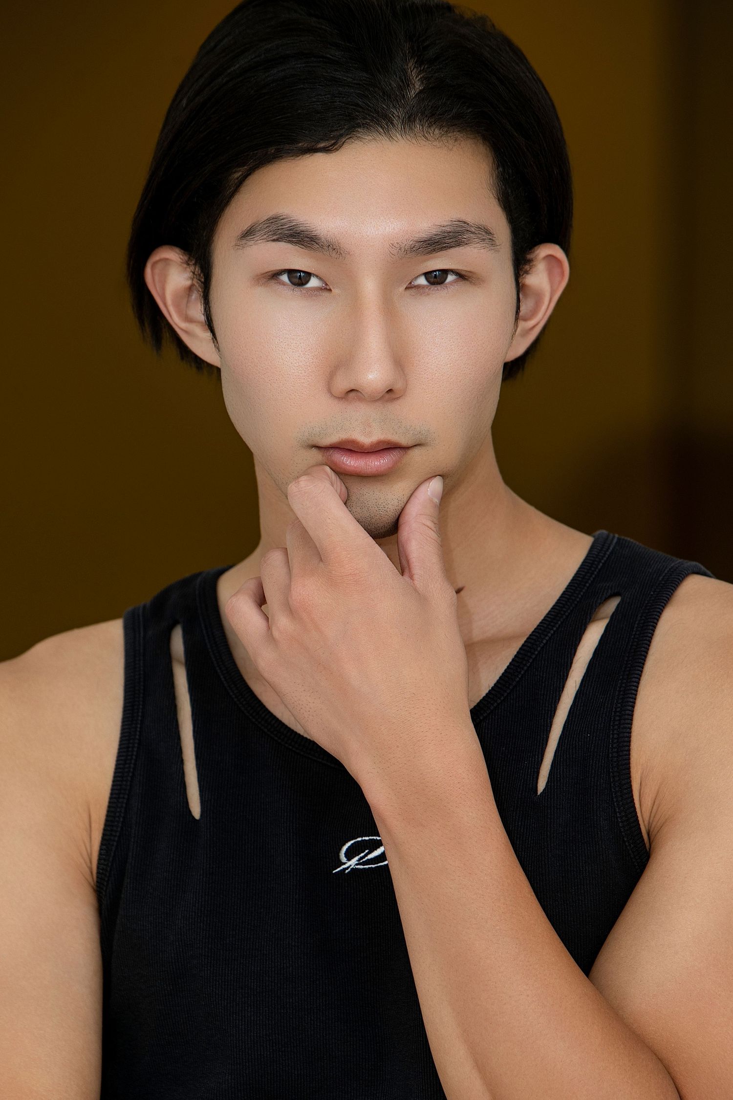 Young Asian man in black sleeveless top touching chin thoughtfully, close-up portrait.
