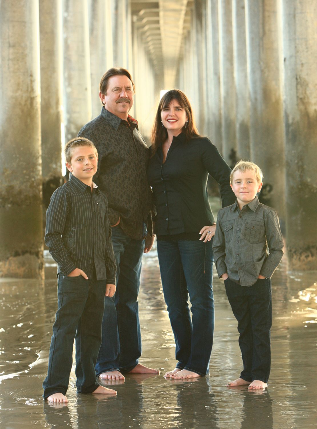 Family of four standing barefoot under a pier on a reflective wet beach at golden hour.