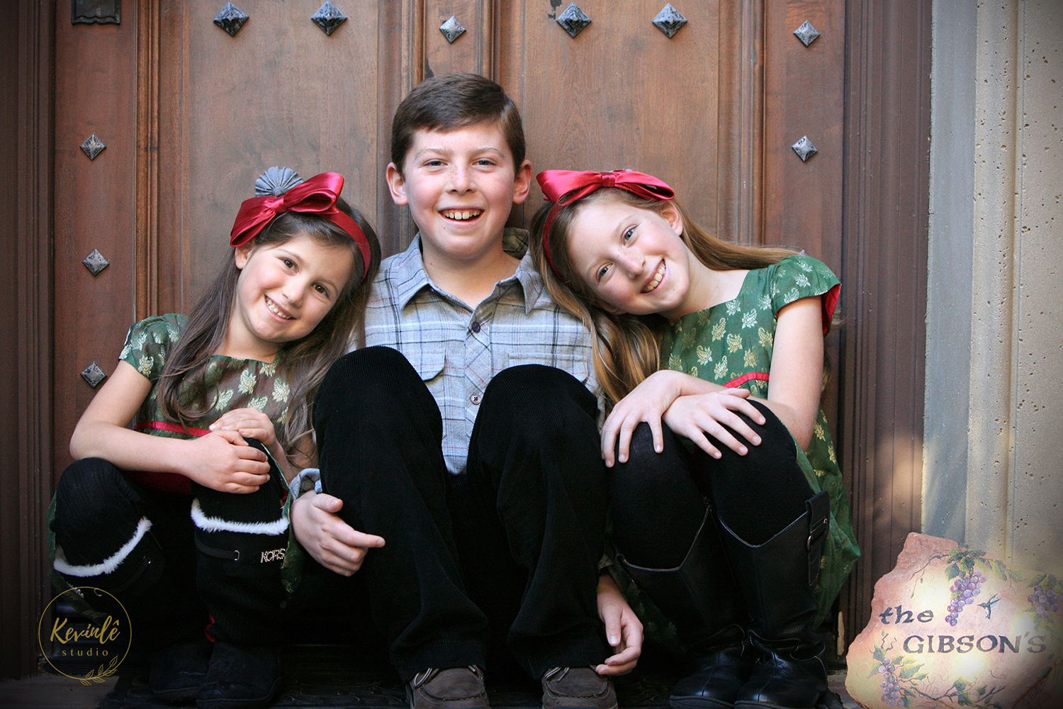 Three siblings sitting together and smiling in front of a wooden door in a professional family portrait.