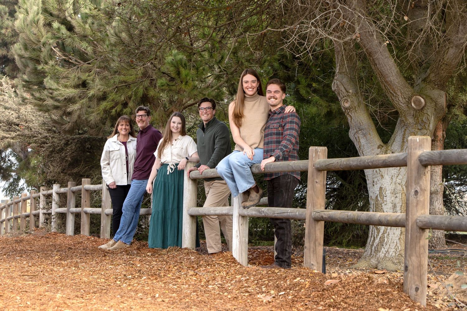 Family of five posing by a rustic wooden fence surrounded by trees on a scenic outdoor trail.